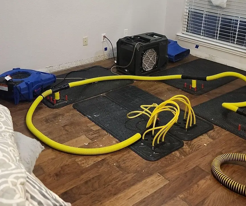 Water restoration equipment with drying mats, hoses, and air movers set up on a wood-look floor inside a home.