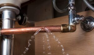 Close-up of clean water spraying from a burst copper supply line beneath a residential kitchen sink with shutoff valve and plumbing connections visible.